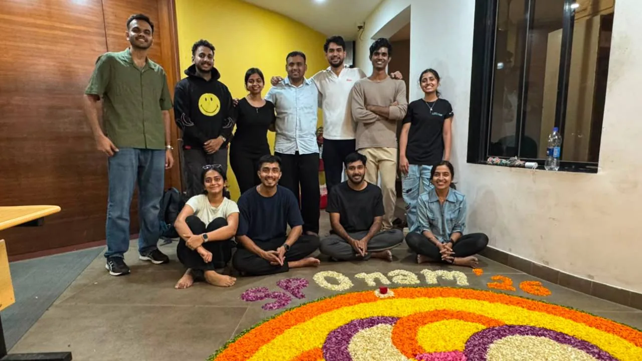 Group of ten people posing indoors beside a large colorful flower rangoli on the floor, celebrating Onam with the text 'Onam 2023' visible.
