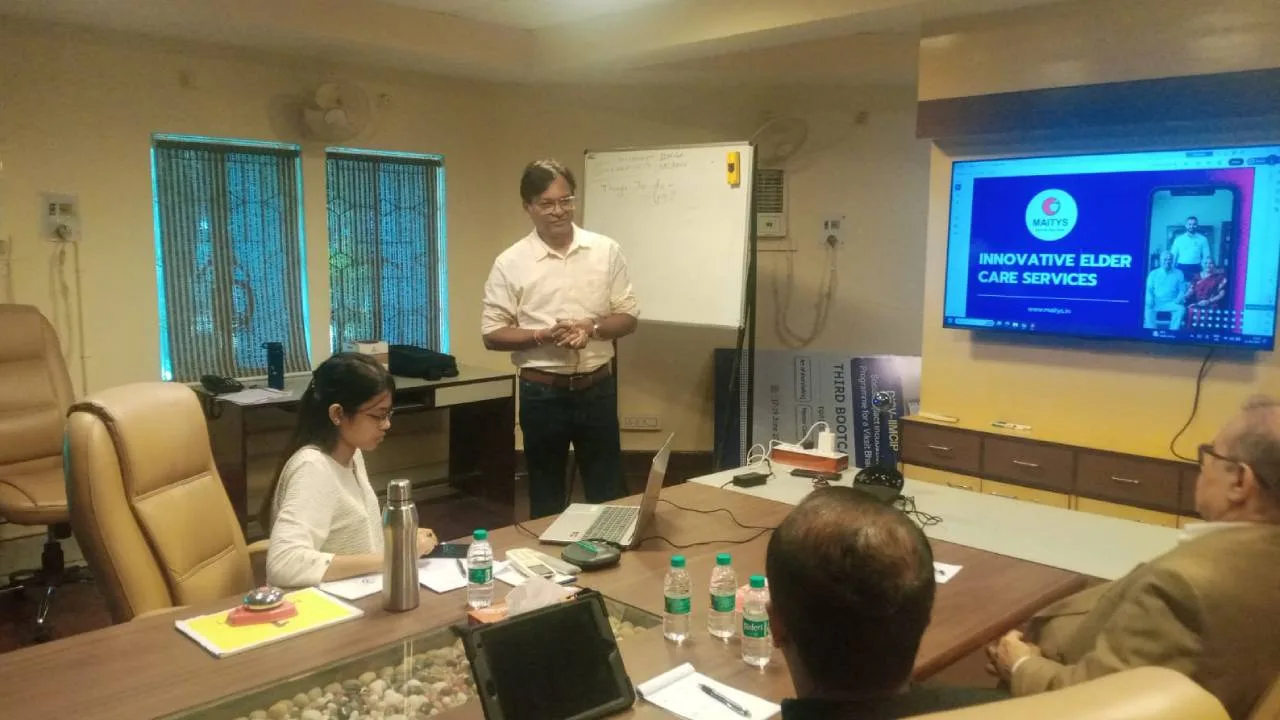 Presenter stands at head of conference table while team watches a slide on a screen: Innovative elder care services, whiteboard in background.
